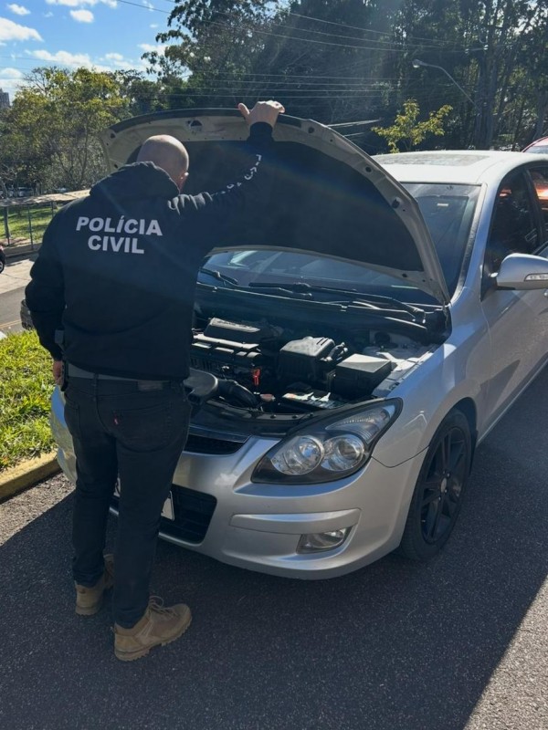 Policial civil realizando verificação de automóvel cinza, com capô aberto.