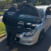 Policial civil realizando verificação de automóvel cinza, com capô aberto.