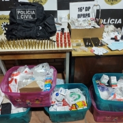 Materiais apreendidos expostos em mesa e ao chão com banner da Polícia Civil