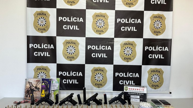 Armas, munições e demais apetrechos apreendidos dispostos em uma mesa com banner da Polícia Civil ao fundo