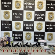 Armas, munições e demais apetrechos apreendidos dispostos em uma mesa com banner da Polícia Civil ao fundo