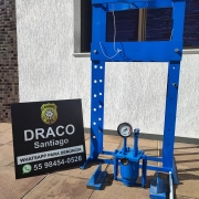 Prensa hidráulica na cor azul, ao lado de placa da Draco de Santiago da Polícia Civil, com fundo em parede branca