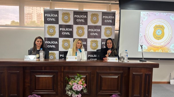 Três mulheres sentadas atrás de uma mesa grande e ao fundo banner da Polícia Civil
