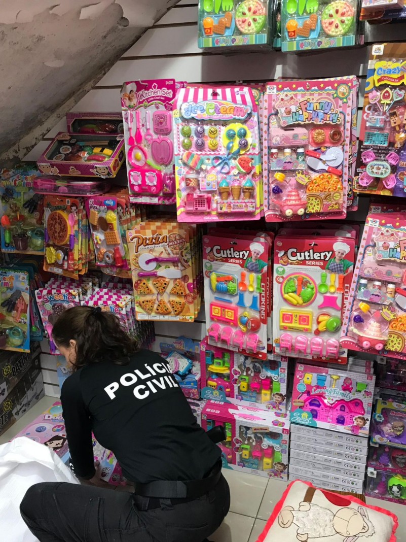 Policial civil abaixada com camiseta preta inscrito "polícia civil" verificando diversos brinquedos coloridos ao chão e pendurados em uma parede