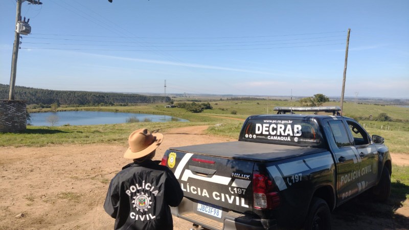 Policial civil de costas usando chapéu, tendo ao lado uma caminhonete viatura com dizeres: Polícia Civil e Debrab e ao fundo campo aberto com vista do horizonte e céu azul