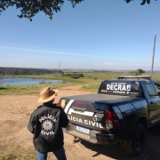 Policial civil de costas usando chapéu, tendo ao lado uma caminhonete viatura com dizeres: Polícia Civil e Debrab e ao fundo campo aberto com vista do horizonte e céu azul