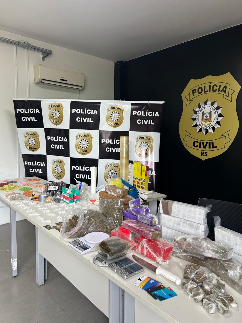 Drogas e demais materiais apreendidos dispostos em mesa com banner da Polícia Civil ao fundo e logo da polícia civil pintado em parede ao fundo
