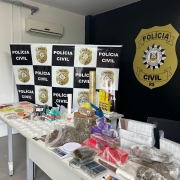 Drogas e demais materiais apreendidos dispostos em mesa com banner da Polícia Civil ao fundo e logo da polícia civil pintado em parede ao fundo