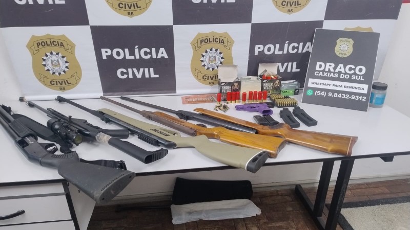 Diferentes armas de fogo e outros materiais como munições expostos em uma mesa com banner da Polícia Civil ao fundo e placa da Draco de Caxias do Sul ao lado