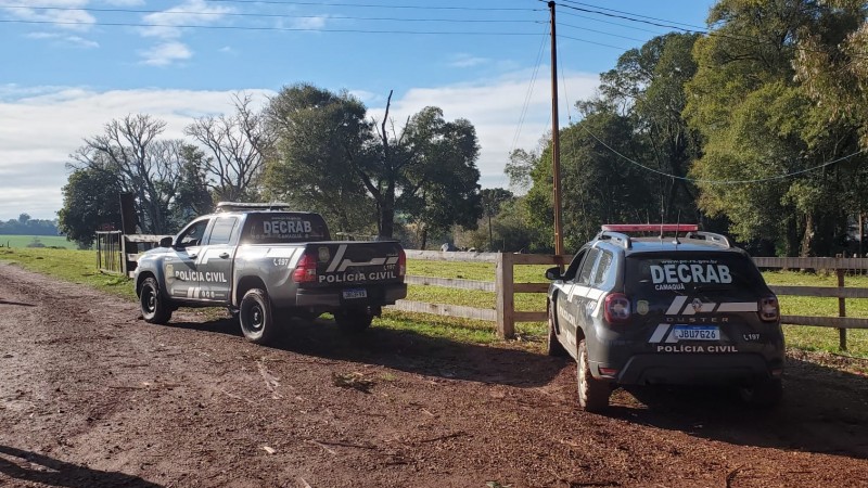Viaturas ostensivas da Polícia Civil, com inscrições da DECRAB, paradas em uma estrada de chão batido em frente a uma porteira de fazenda