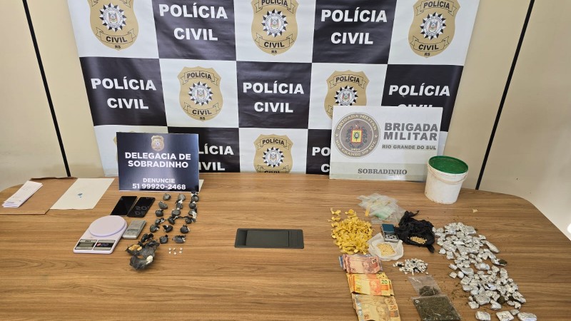 Drogas e apetrechos apreendidos dispostos em cima de uma mesa com banner da Polícia Civil ao fundo, em conjunto com o da Brigada Militar