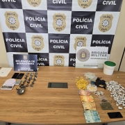 Drogas e apetrechos apreendidos dispostos em cima de uma mesa com banner da Polícia Civil ao fundo, em conjunto com o da Brigada Militar