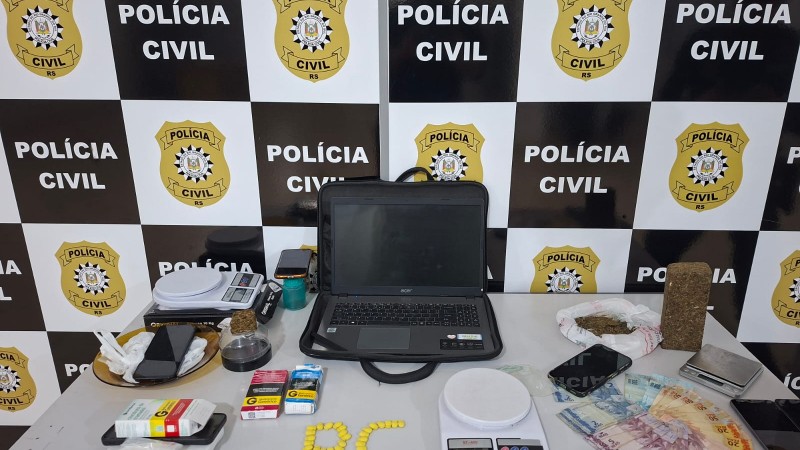Diferentes itens como drogas, balanças, computador, medicamentos e outros materiais expostos em uma mesa com banners preto e branco da Polícia Civil ao fundo