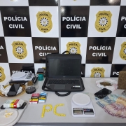 Diferentes itens como drogas, balanças, computador, medicamentos e outros materiais expostos em uma mesa com banners preto e branco da Polícia Civil ao fundo
