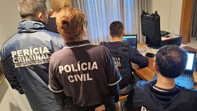 Policiais civis e peritos do IGP em pé e sentados com computadores em uma residência