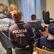Policiais civis e peritos do IGP em pé e sentados com computadores em uma residência