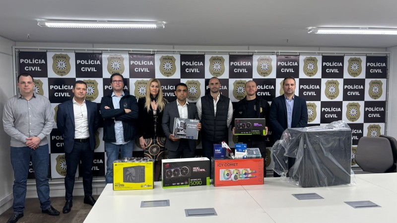 Pessoas em pé posadas para foto com banners da Polícia Civil ao fundo e equipamentos eletrônicos alocados em cima de uma mesa