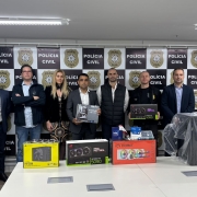 Pessoas em pé posadas para foto com banners da Polícia Civil ao fundo e equipamentos eletrônicos alocados em cima de uma mesa
