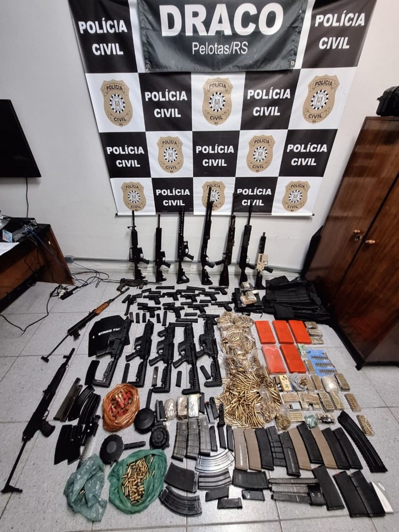 Armas e munições dispostos ao chão com banner da Polícia Civil ao fundo