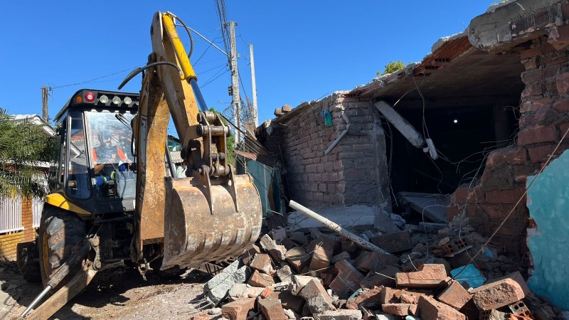 Retroescavadeira amarela destruindo construção fortificada, com tijolos caídos e partes de grossas paredes parcialmente destruídas