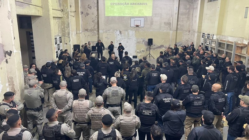 Policiais civis e militares em pé olhando projeção em parede sobre a operação