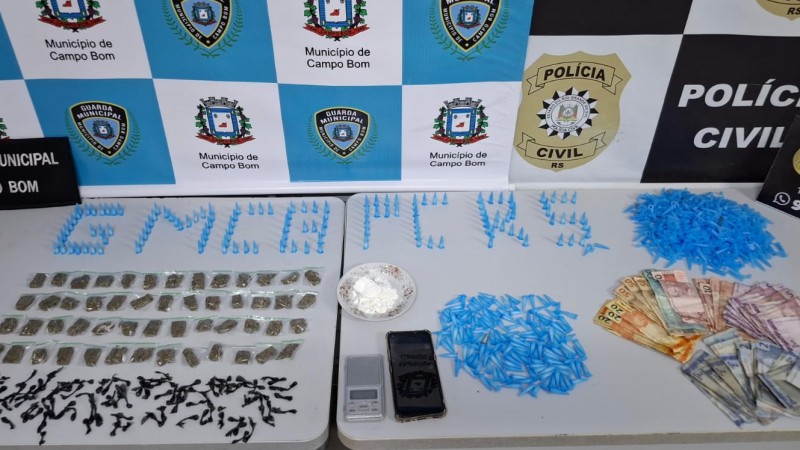 Pinos, entorpecentes, dinheiro e outros materiais apreendidos dispostos sobre uma mesa branca, com banners da Polícia Civil e Guarda Municipal de Campo Bom ao fundo