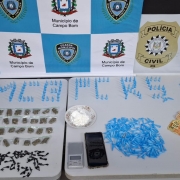 Pinos, entorpecentes, dinheiro e outros materiais apreendidos dispostos sobre uma mesa branca, com banners da Polícia Civil e Guarda Municipal de Campo Bom ao fundo