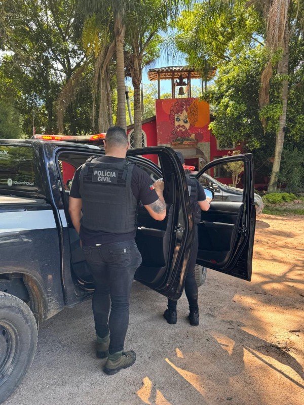 Policiais com portas de viatura abertas, vestindo roupas prestas e um homem de costas com colete inscrito: Polícia Civil. 