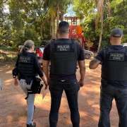 Três policiais de costas, sendo dois homens e mulher à esquerda, com coleta com a inscrita: Polícia Civil