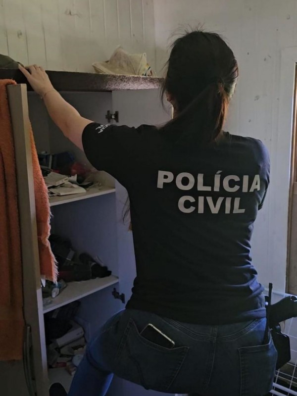 Policial civil mulher de costas, com camiseta preta com inscrições: Polícia Civil, fiscalizando residência