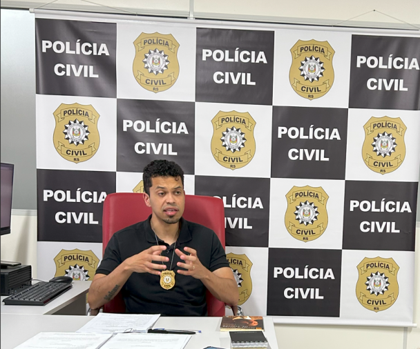 Homem pardo sentado em uma cadeira vermelha com um computador à esquerda e banner da Polícia Civil ao fundo