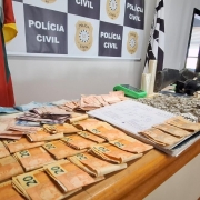 C&eacute;dulas de dinheiro, drogas e demais itens expostos em uma mesa, tendo o banner da Pol&iacute;cia Civil ao fundo