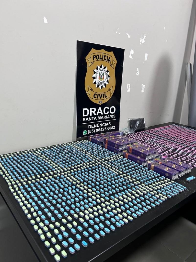 Comprimidos de ecstasy de diversas cores espalhados sobre uma mesa com placa da Pol&iacute;cia Civil com inscri&ccedil;&atilde;o Draco ao fundo