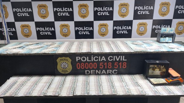 Diversos c&eacute;dulas de reais espalhadas sobre mesas e ao fundo banner da Pol&iacute;cia Civil