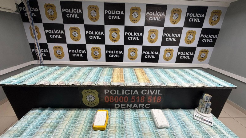C&eacute;dulas de 100 e 200 reais dispostas em cima de mesas com banner da pol&iacute;cia civil ao fundo