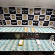 C&eacute;dulas de 100 e 200 reais dispostas em cima de mesas com banner da pol&iacute;cia civil ao fundo
