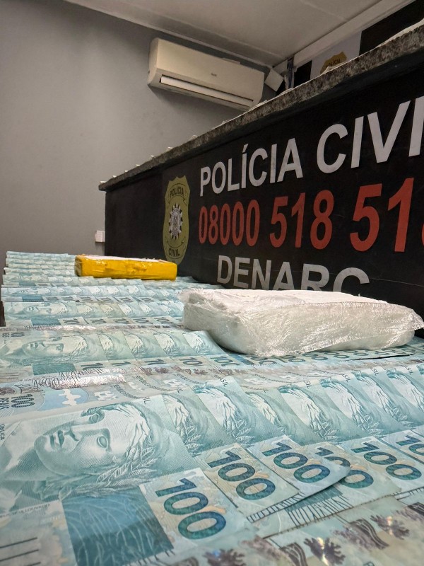 Cédulas de 100 reais em primeiro plano com dois pacotes branco e amarelo contendo cocaína dispostos em uma mesa