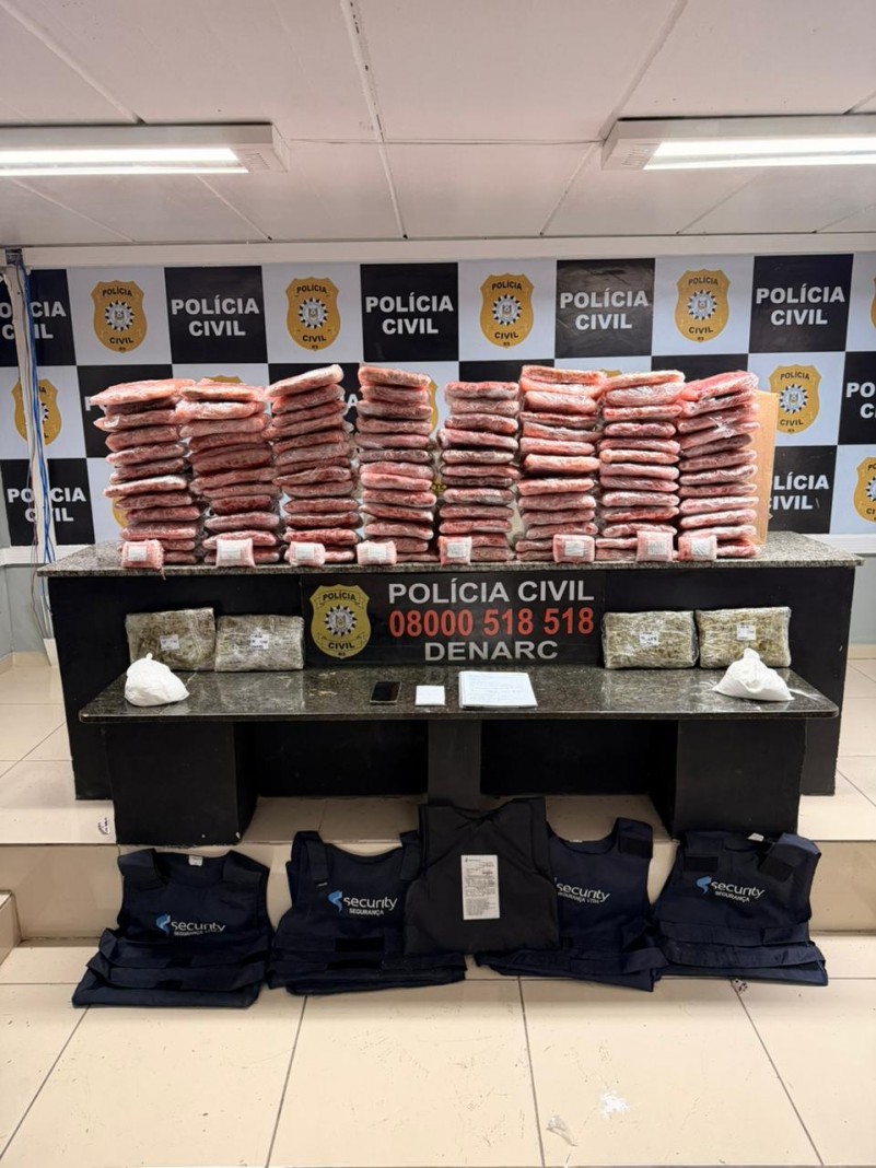 Pacotes de cor avermelhada dispostos em cima de mesa com banner da pol&iacute;cia civil ao fundo, al&eacute;m de coletes bal&iacute;sticos e outros materiais apreendidos