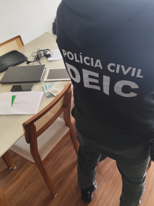 Policial de costas com dizeres Pol&iacute;cia Civil Deic no casaco e computadores, folhas e eletr&ocirc;nicos em mesa ao fundo