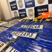 Diversas placas veiculares, equipamentos de inform&aacute;tica e outros materiais dispostos em mesa com banner da Pol&iacute;cia Civil ao fundo