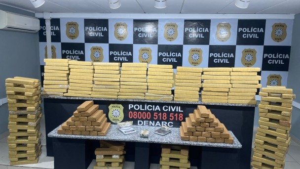 Centenas de tabletes de drogas embalados na cor amarelo dispostos sobre bancadas e no ch&atilde;o com banner da Pol&iacute;cia Civil ao fundo e no centro placa com os dizeres: Pol&iacute;cia Civil - 0800 518 518 - Denarc
