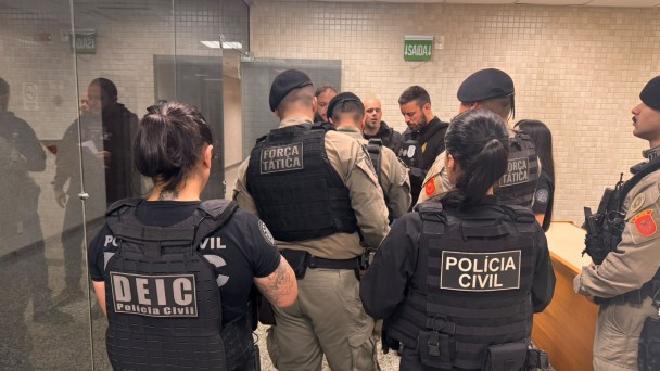 Diversos policiais civis e militares, entre homens e mulheres, conversando em uma sala com parede de vidro &agrave; esquerda