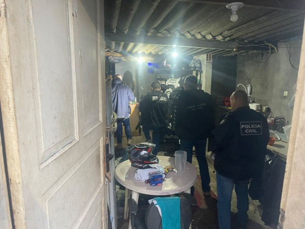 Diversos policiais homens vestidos com roupas e coletes da Polícia Civil, roupas e equipamentos majoritariamente pretos, dentro de um casebre 