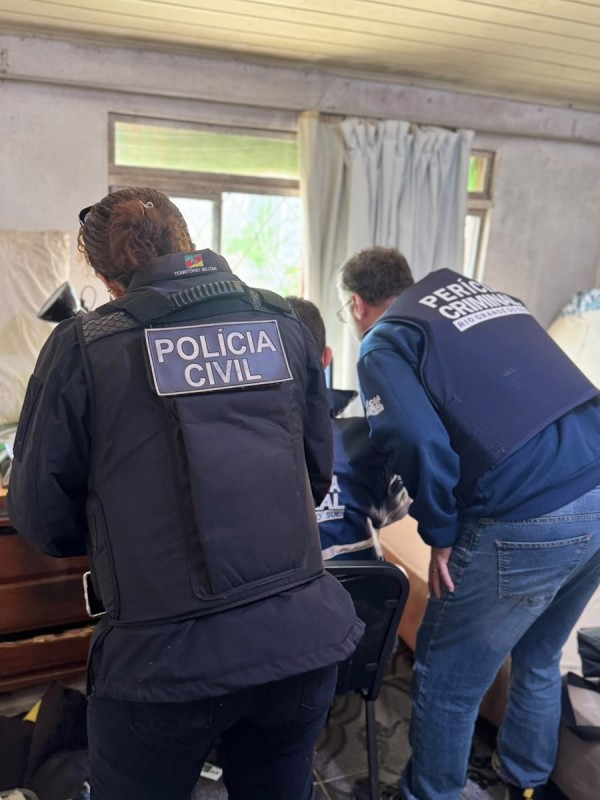 Policial civil mulher e perito criminal homem de costas observando o que deduz-se ser algum equipamento eletr&ocirc;nico, mas n&atilde;o vis&iacute;vel na imagem, dentro de uma esp&eacute;cie de sala ou quarto
