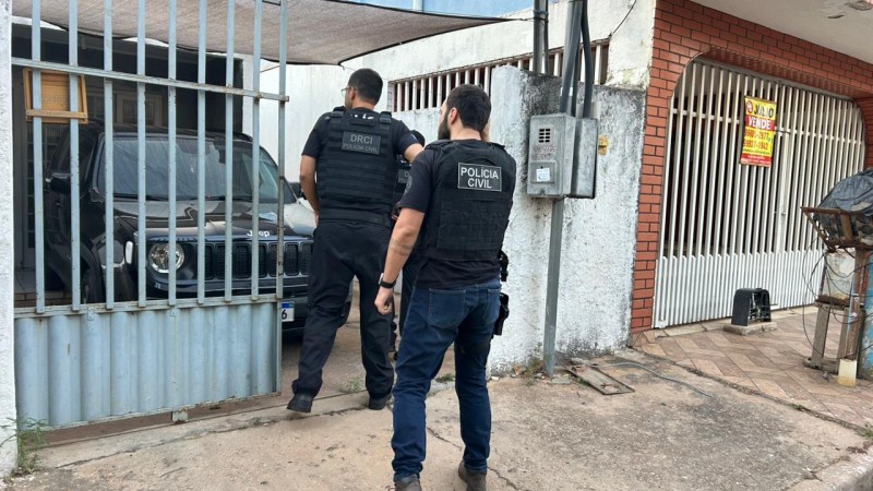 Dois policiais civis homens vestindo roupas e colete pretos entrando em resid&ecirc;ncia com port&atilde;o semiaberto e ve&iacute;culo preto na garagem