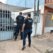 Dois policiais civis homens vestindo roupas e colete pretos entrando em resid&ecirc;ncia com port&atilde;o semiaberto e ve&iacute;culo preto na garagem