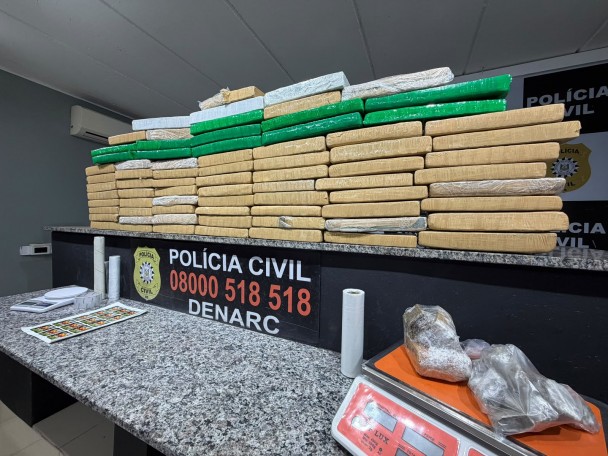 Diversos tabletes de drogas embalados em coloração amarela e verde, além de outros materiais citados no texto da matéria expostos em bancadas, tendo ao centro escrita: Polícia Civil - 0800 518 518 - Denarc