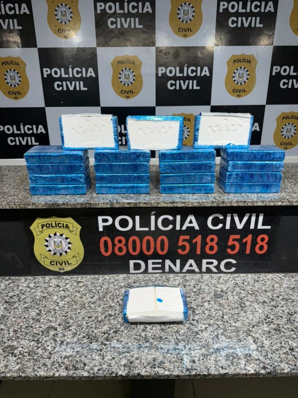 Diversos tabletes de drogas branca embalados em coloração azul, expostos em bancadas com banner da Polícia Civil ao fundo e ao centro escrita: Polícia Civil - 0800 518 518 - Denarc