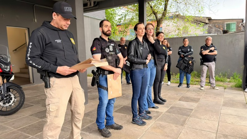 Policiais civis homens e mulheres em p&eacute; em linha com pastas nas m&atilde;os embaixo de &aacute;rea coberta falando para p&uacute;blico n&atilde;o aparente na imagem