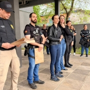 Policiais civis homens e mulheres em p&eacute; em linha com pastas nas m&atilde;os embaixo de &aacute;rea coberta falando para p&uacute;blico n&atilde;o aparente na imagem